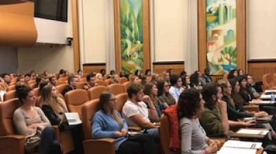 Audience seated in rows of tan chairs attentively listens to a presentation in a large room with decorative wall art and high ceilings.