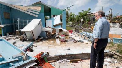 Man surveys damage to a house with collapsed walls and scattered debris after a storm.