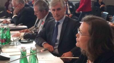 People in formal attire seated at a conference table with microphones and documents; green water bottles are on the table.
