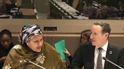 Two people are seated and engaged in discussion at a conference table. One person wears a patterned headscarf and the other is in a suit and tie. Other attendees are visible in the background.