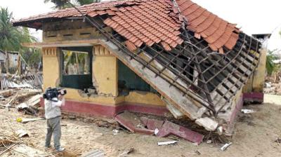 A person films a partially collapsed house with a damaged roof, surrounded by debris, under a clear sky.