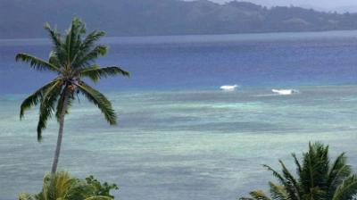 Tropical beach scene with palm trees, turquoise water, and distant hills under a cloudy sky.