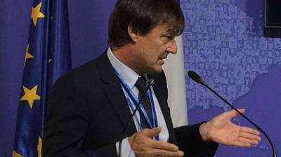A man in a suit speaks at a podium with microphones, standing in front of an EU flag and a blue background.
