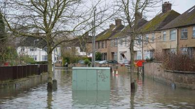 Flooded residential street with submerged road, lined with houses and bare trees. Barricade and traffic cones partially visible. Overcast sky above.