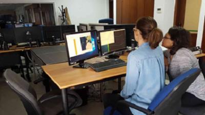 Two people in an office sit at a desk with dual monitors displaying maps and data. Multiple empty chairs and more computers are in the background.