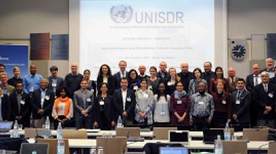 A diverse group of people poses in front of a presentation screen displaying the UNISDR logo at a conference room.