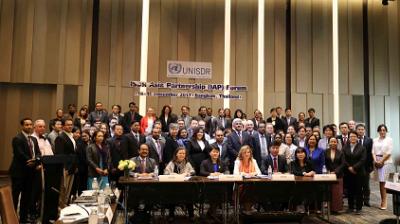 A large group of people in formal attire pose for a group photo at the UNISDR Asia Partnership Forum in a conference room with a sign reading "Bangkok, Thailand" in the background.