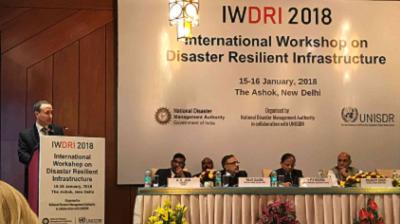 A speaker addresses attendees at the IWDRI 2018 International Workshop on Disaster Resilient Infrastructure in New Delhi, with panelists seated at a table.