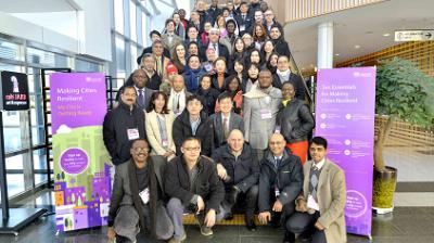 A large group of people poses for a photo on a staircase at an indoor conference, flanked by banners about making cities resilient.
