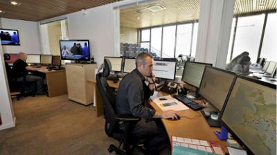 A man in an office setting is using a phone while seated at a desk with multiple computer monitors. Other workstations and personnel are visible in the background.