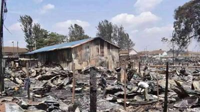 A corrugated metal building stands amidst rubble and debris, with a few people walking around. Burnt, charred remains are scattered throughout the area, and trees are visible in the background.