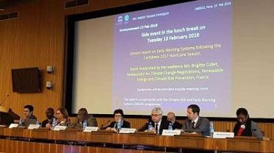 A panel of nine people sit at a long table with microphones during a side event at a conference, with a large presentation screen behind them displaying event details.