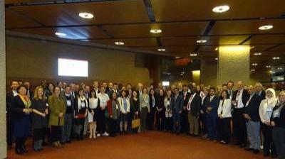 A large group of people stands indoors in a conference or event hall, posing for a group photo under a high ceiling with lights.