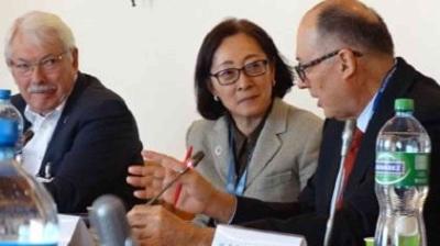 Three people in business attire sit at a conference table with microphones, engaged in discussion. Bottles of water are on the table.