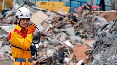 A rescue worker in a yellow uniform and helmet communicates on a radio while standing in front of a large pile of rubble.