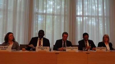 Five people sit behind a desk in a conference room, with nameplates in front of them and large windows with curtains in the background.