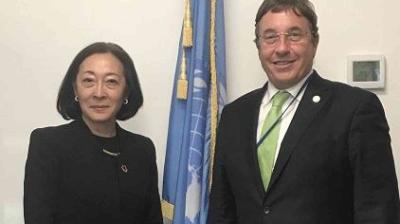 Two people in formal attire stand in front of a United Nations flag, posing for a photo indoors.