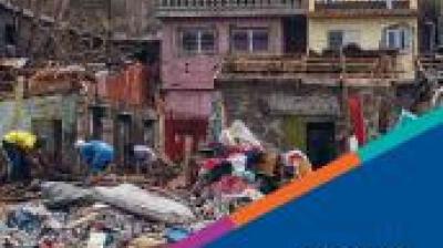 People search through debris in front of damaged buildings; piles of rubble and destroyed materials are visible.