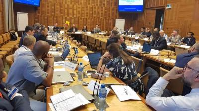 A group of people in business attire sit around a large conference table with laptops, papers, and microphones in a wood-paneled meeting room.