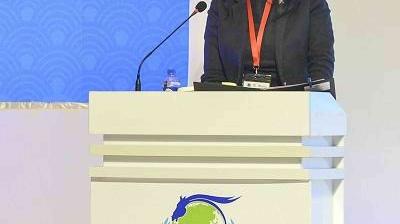 Person speaking at a podium with a microphone, wearing a black jacket and red lanyard. The podium features a logo with a horse and globe.