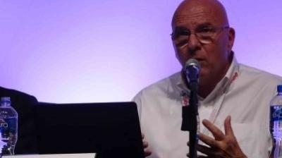 Bald man in glasses speaks into a microphone at a panel discussion, seated at a table with a laptop and water bottles.