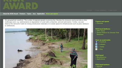 Eroded riverbank with trees and exposed roots; two people walk along the edge, one in colorful clothing, under a cloudy sky.