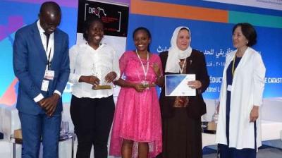 A group of five people stands on a stage. Two women hold trophies, and another holds a certificate. A screen and banner are in the background.