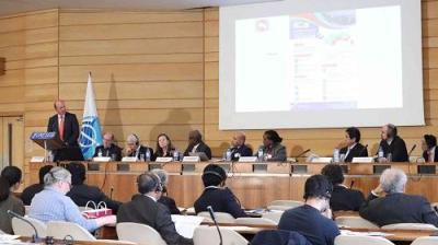 A panel of nine speakers sit at a long table, presenting to an audience. A large screen displays information above them. The event takes place in a formal conference room.