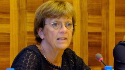 A woman with short brown hair and glasses speaks into a microphone at a wooden-panel table.