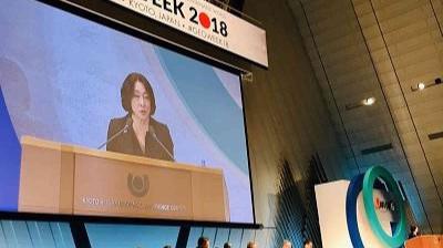 A person speaks at a podium on a stage during an event named "OECD WEEK 2018" in Kyoto, Japan. Several people are seated at a table beside the speaker.