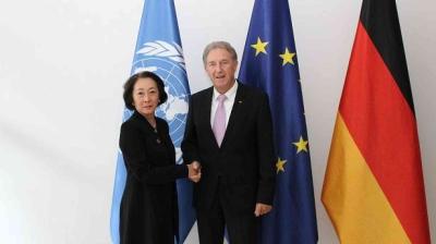 Two individuals shake hands in front of United Nations, European Union, and German flags.