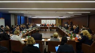 A large group of people seated around a U-shaped conference table in a meeting room with banners at the far end.