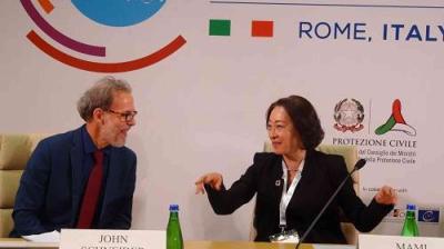 Two people sit at a conference table in front of a backdrop with logos and "Rome, Italy" text, engaged in conversation. Bottled water and nameplates are visible.