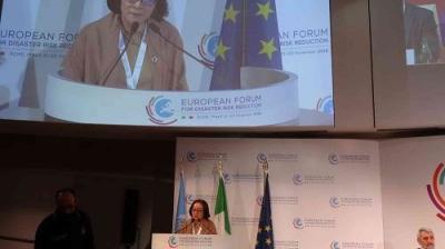 A person speaks at a podium with logos for the European Forum for Disaster Risk Reduction. Flags and a large screen display are in the background.