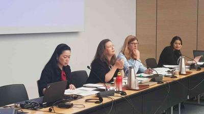 Four people sit at a conference table with laptops and documents, engaged in discussion.