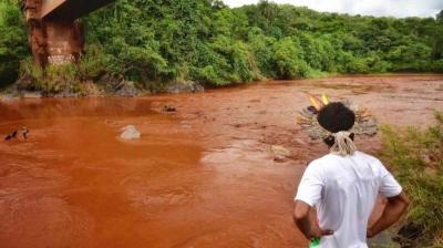 A person wearing a feathered headdress looks at a reddish-brown river beneath a bridge, with dense green vegetation on both banks.