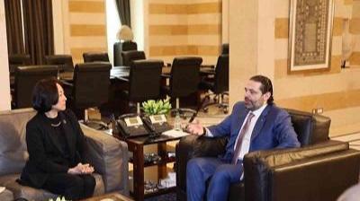 Two people sit across from each other in an office lounge area, engaged in conversation. Office chairs and tables are visible in the background.