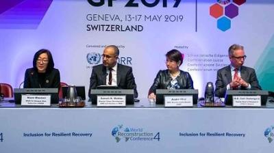 Four individuals are seated at a panel discussion table with microphones in front of them. A banner behind them reads "GP 2019, Geneva, Switzerland.