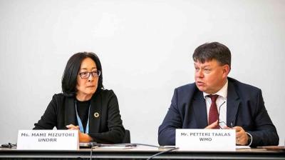 Two people are seated at a table with nameplates, having a discussion. The woman on the left has dark hair and glasses, while the man on the right is wearing a suit and tie.