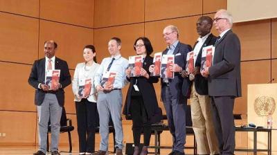 Seven people stand in a row on stage holding documents, in front of a podium with a United Nations emblem in a wood-paneled room.