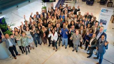 A large group of people in business attire stand together indoors, smiling and waving at the camera.