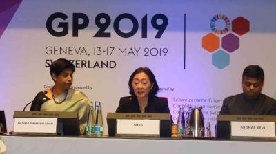 Three individuals sit at a conference table with microphones. A banner behind them reads "GP 2019 Geneva, 13-17 May 2019 Switzerland.