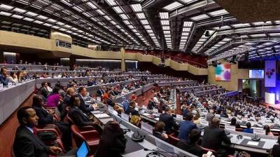 A large conference hall filled with attendees seated in rows, facing a presentation on stage. The ceiling features a grid of lights.