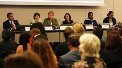 A panel of six people sits at a long table in front of an audience, engaged in a discussion. They are positioned in a conference or meeting setting.
