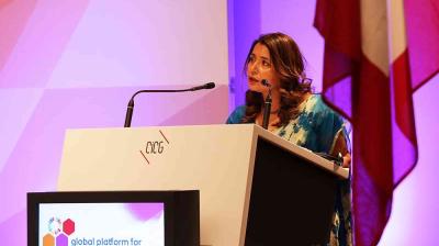 A woman stands at a podium labeled "CICG," speaking into a microphone at a conference with a display screen and flags in the background.