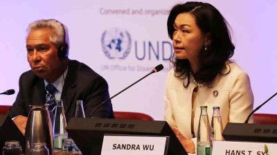 Two individuals sit at a conference panel with microphones, water bottles, and nameplates. A UN logo is visible in the background.
