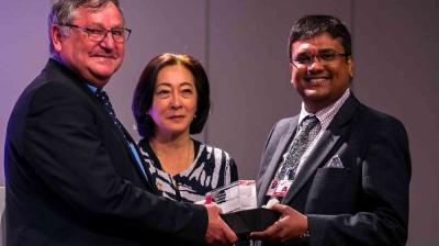 Three people in formal attire stand together; two men shake hands while holding a boxed item, with a woman standing between them, indoors against a plain background.