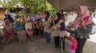 A group of women and children gather outdoors under a canopy, with some standing and speaking while others sit and listen.
