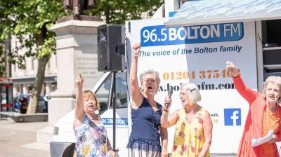 Four women standing and speaking into a microphone near a Bolton FM vehicle. One woman raises her fist, and a statue is visible in the background.