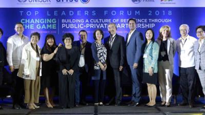 A group of people in business attire stands on stage in front of a blue banner that reads "Top Leaders Forum 2018: Building a Culture of Resilience Through Public-Private Partnership.